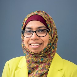 Mahfuja Malik is wearing black glasses, a multi-colored printed head scarf and a bright yellow blazer. She is smiling.